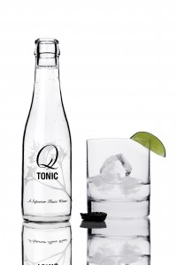 Q-Tonic bottle with glass Q Tonic bottle with rocks glass partially filled with ice and a small amount of gin. A lime wedge is on the rim of the glass and the black Q Tonic bottle cap is off the bottle, upside down, next to the rocks glass.