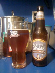 Samuel Adams Dunkelweizen poured into a Samuel Adams glass next to the bottle. The beer is the same color as honey in the background.