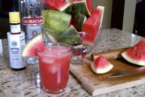 Fresh cut watermelon, a bottle vodka, and a bottle of bitters behind a glass 