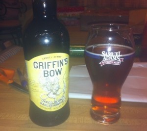 A bottle of Samuel Adams Griffin's Bow next to a Samuel Adams beer glass filled with amber Griffin's Bow
