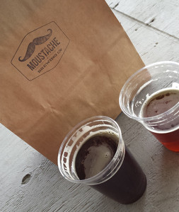 Two plastic pints of Moustache Brewing beer, one amber and one dark, by a paper bag with Moustache’s logo