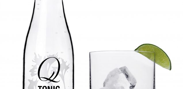 Q Tonic bottle with rocks glass partially filled with ice and a small amount of gin. A lime wedge is on the rim of the glass and the black Q Tonic bottle cap is off the bottle, upside down, next to the rocks glass.