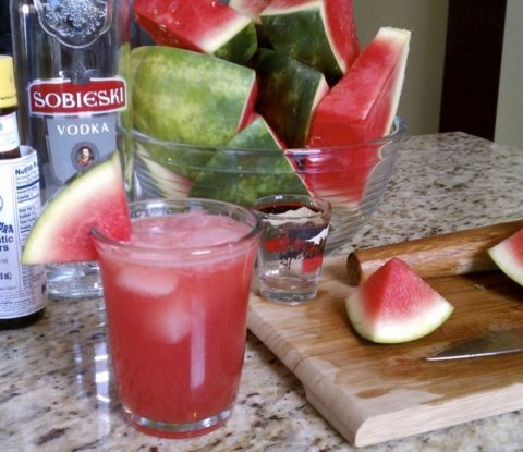 Fresh cut watermelon, a bottle vodka, and a bottle of bitters behind a glass
