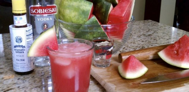 Fresh cut watermelon, a bottle vodka, and a bottle of bitters behind a glass