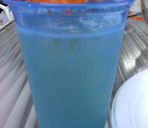 A blue plastic cup filled with a Lillet spritzer. An orange paper umbrella is hanging over the rim.