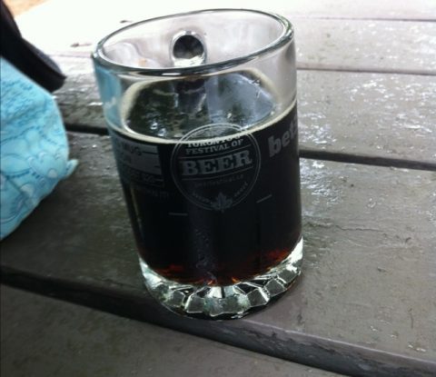 A Toronto’s Festival of Beer glass mug partly filled with Krombacher Dark on a wooden picnic table.