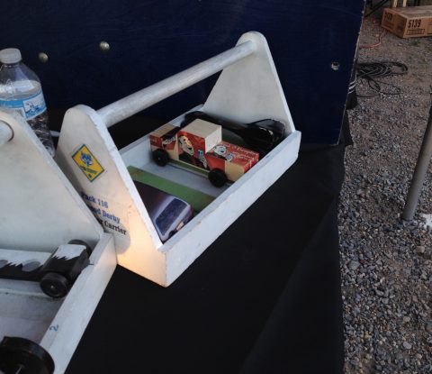 Several pinewood derby cars wait in carriers to be placed in the next race. Our Drunk and Unemployed car with stickers on a plain pinewood truck, did not make it to the next round.
