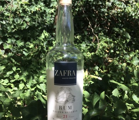 A golden moth sits atop a bottle of Zafra Master Reserve. The bottle is half empty, at it stands amongst a background of green ivy.