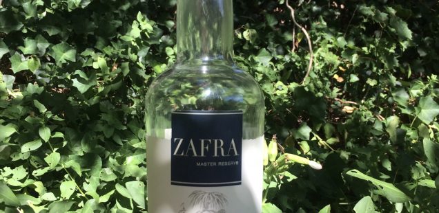 A golden moth sits atop a bottle of Zafra Master Reserve. The bottle is half empty, at it stands amongst a background of green ivy.