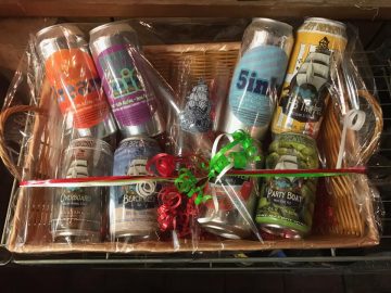 Eight cans of Port Jefferson Brewing beer and a pint glass with Port Jefferson Brewing's logo in a wrapped gift basket.