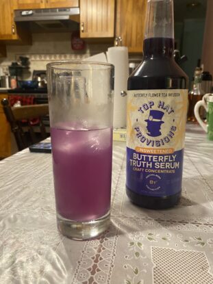 A half of a highball glass of Faux Paloma, a lilac-colored cold drink, next to a bottle of Top Hat Provisions Butterfly Truth Serum