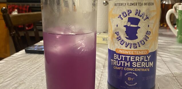 A half of a highball glass of Faux Paloma, a lilac-colored cold drink, next to a bottle of Top Hat Provisions Butterfly Truth Serum