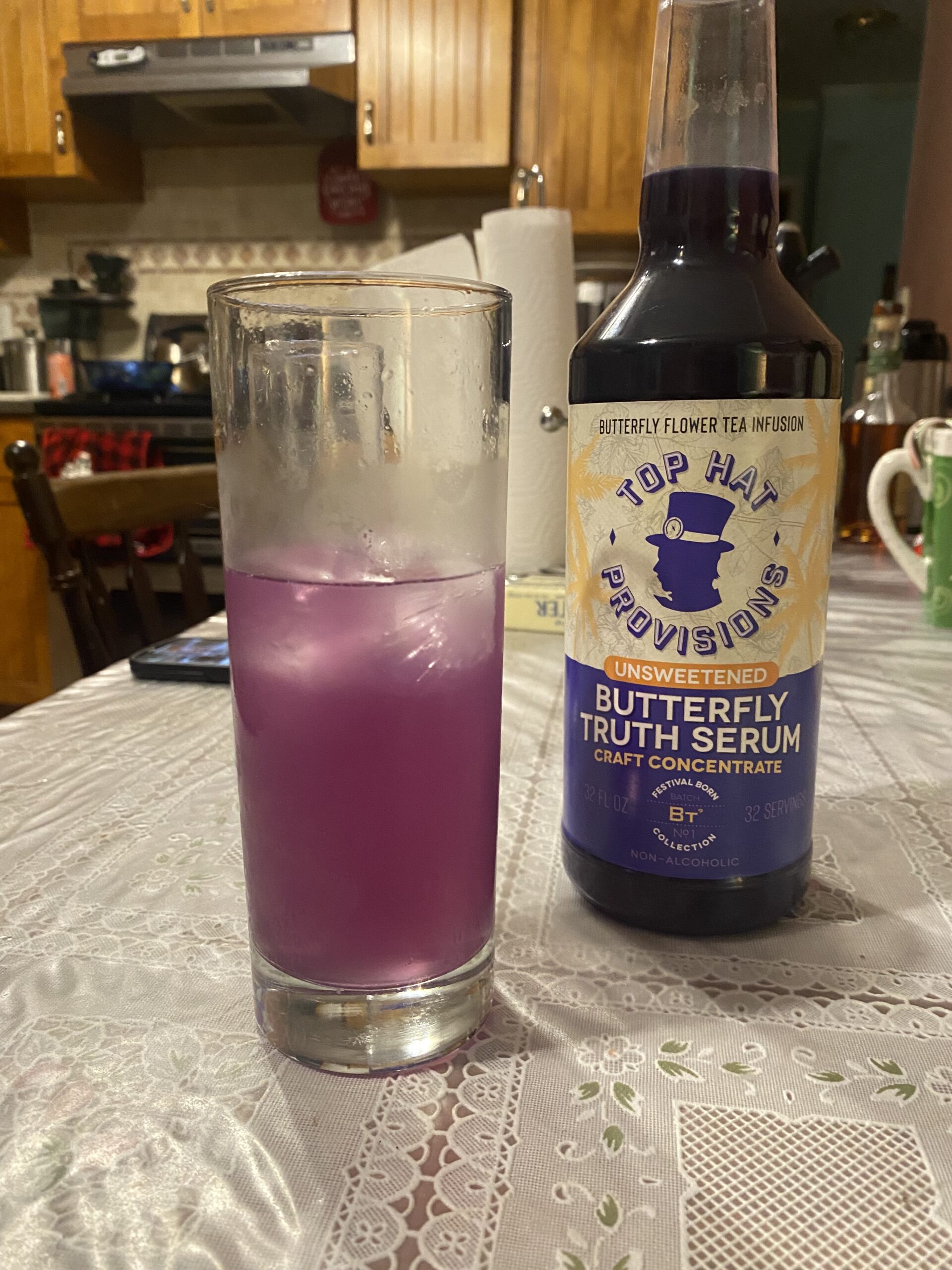 A half of a highball glass of Faux Paloma, a lilac-colored cold drink, next to a bottle of Top Hat Provisions Butterfly Truth Serum