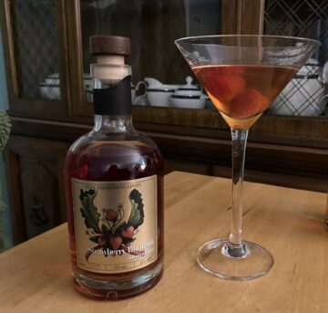 A bottle of Strawberry Rhubarb Moonshine next to a martini glass containing a dark orange cocktail with two strawberries