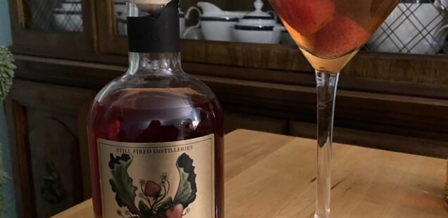 A bottle of Strawberry Rhubarb Moonshine next to a martini glass containing a dark orange cocktail with two strawberries