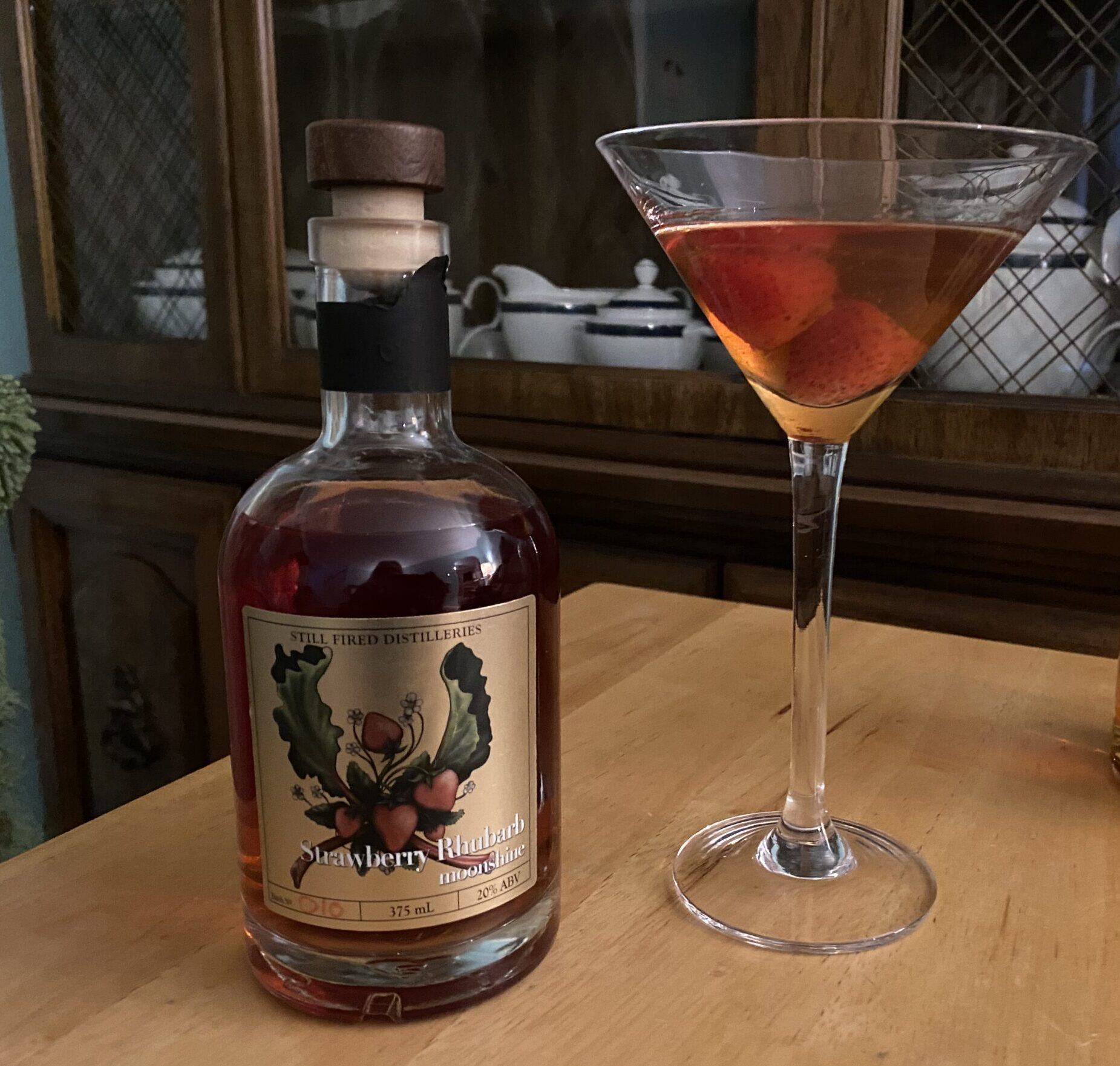 A bottle of Strawberry Rhubarb Moonshine and a cocktail containing two strawberries in a martini glass